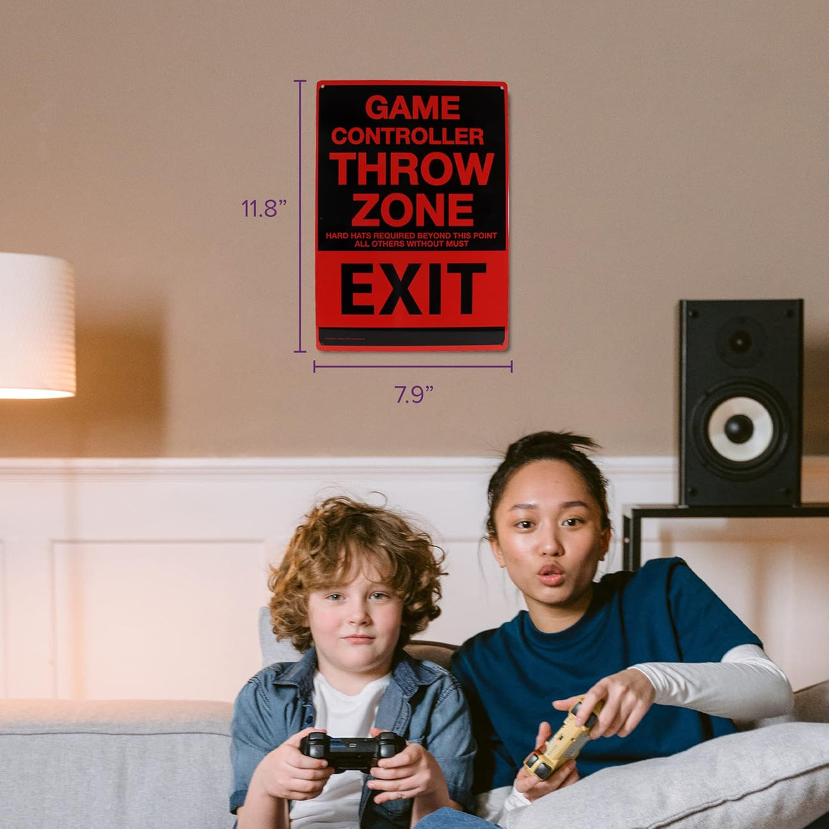 Two children sit on a couch, deeply engaged in their video games, beneath the Metal Tin Sign "Game Controller THROW ZONE" Bedroom Gamer Wall Decor #F-101, known for its humor and precision.