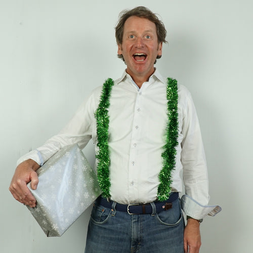 Man wearing 🎄 Tinsel Suspenders 🎅, the ultimate holiday dress accessory, over a white shirt and jeans, holding a silver gift box against a plain background—perfect party bling for Christmas celebrations.