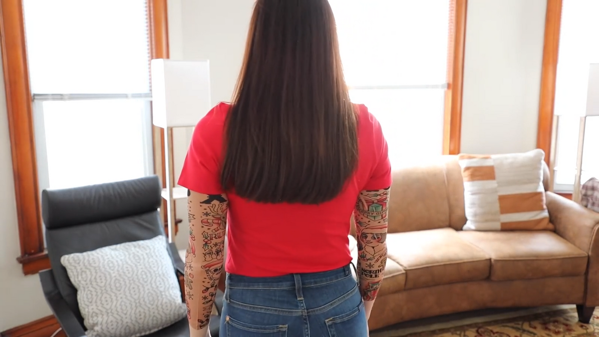 A person with long brown hair, a red shirt, jeans, and visible tattoos wears 🎄 Tinsel Suspenders—sparkling Christmas party bling—in a living room with a sofa, lamp, and armchair.