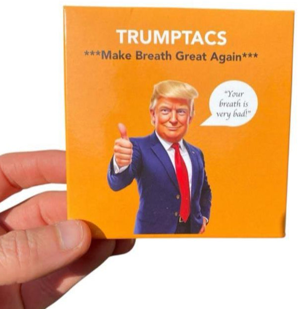 A hand holds a Trumptacs Mints box—funny Donald Trump candy with "Make Breath Great Again" and a cartoon man giving a thumbs up. Hilarious gag gift for fans of Presidential Mints! Box contains 30 mints.