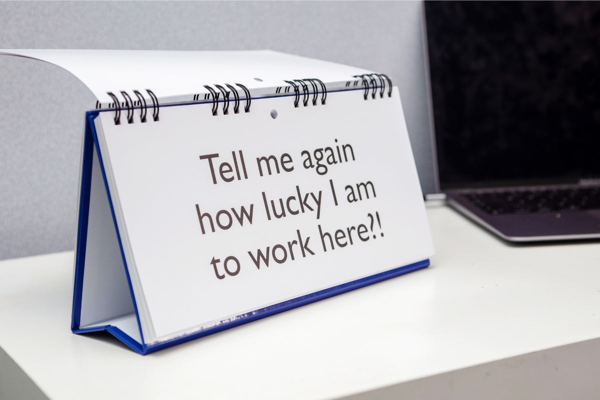 The "Workplace Mood Swings" desk spiral flip book, featuring the humorous message "Tell me again how lucky I am to work here?!", sits next to a laptop on a white table—a perfect gift for co-workers to inspire office chatter.