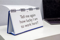 The "Workplace Mood Swings" desk spiral flip book, featuring the humorous message "Tell me again how lucky I am to work here?!", sits next to a laptop on a white table—a perfect gift for co-workers to inspire office chatter.