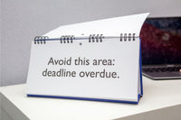 On a desk with a partially visible laptop, a sign from the Workplace Mood Swings - Desk Spiral Flip Book humorously displays "Avoid this area: deadline overdue," hinting at its owner's changing moods.